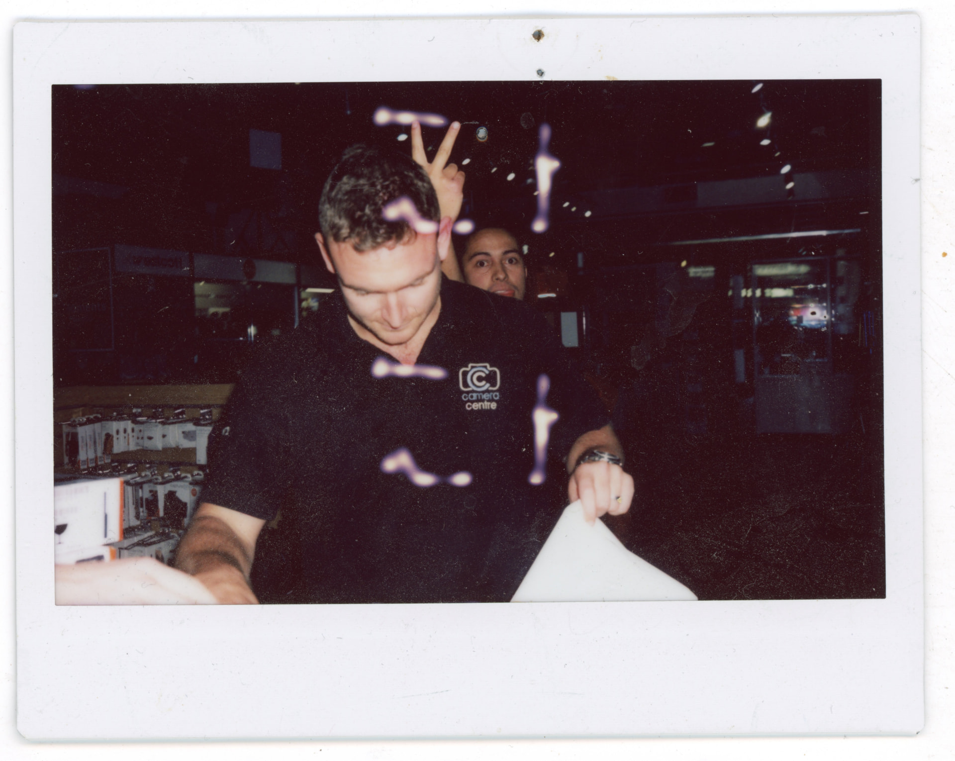 Instax photo of Terry working a Sunday shift in the Camera Centre UK shop while a colleague photobombs with peace signs