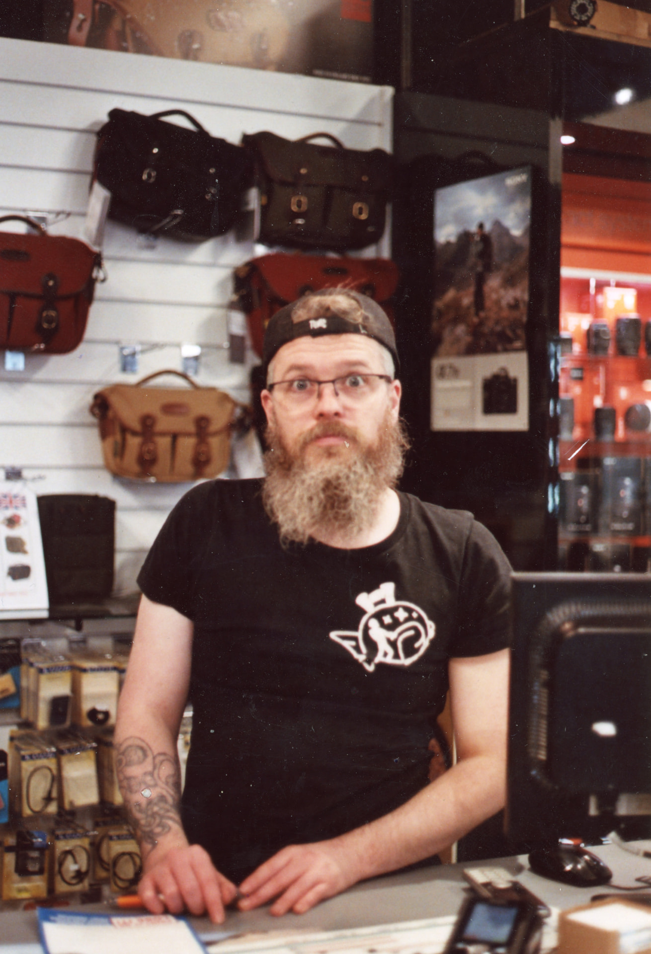 Luke behind the counter at Camera Centre UK, surrounded by camera bags and accessories